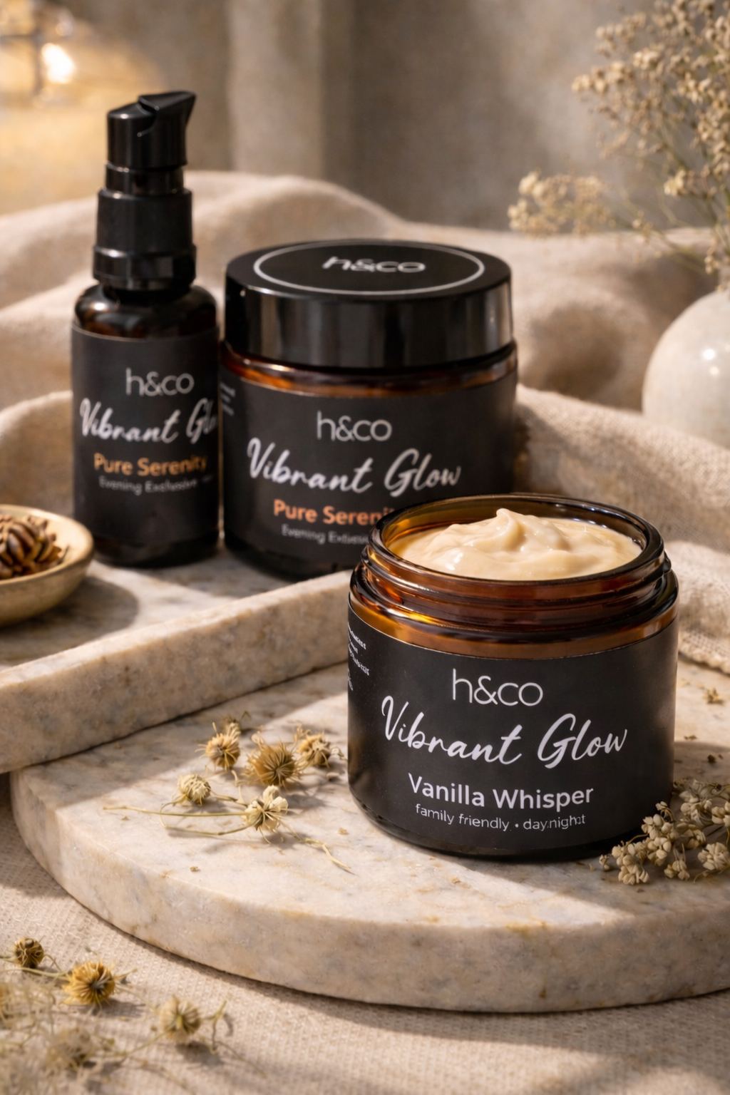 Three h&co Vibrant Glow skincare products in amber glass containers sit on a stone slab surrounded by dried flowers.
