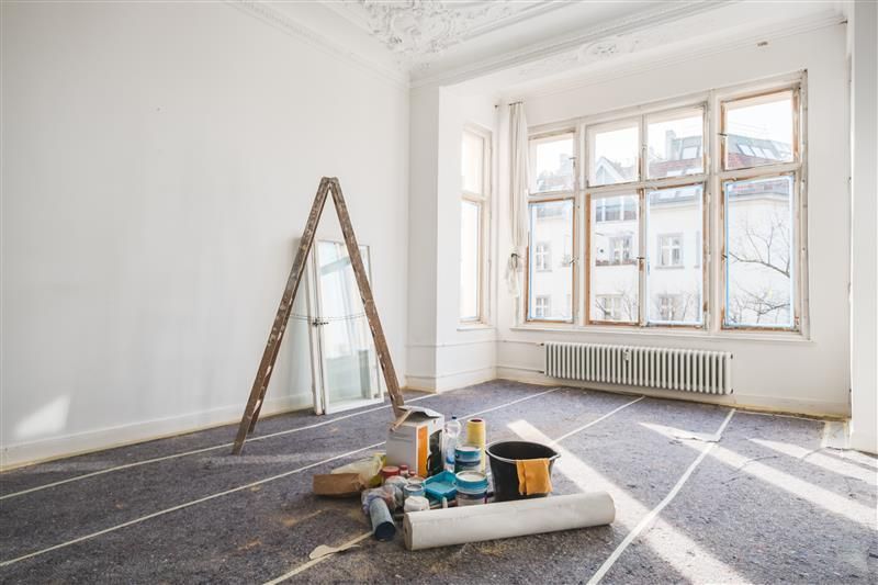 An empty, white-walled room undergoing renovation, featuring a wooden ladder, painting supplies, and rolled wallpaper.