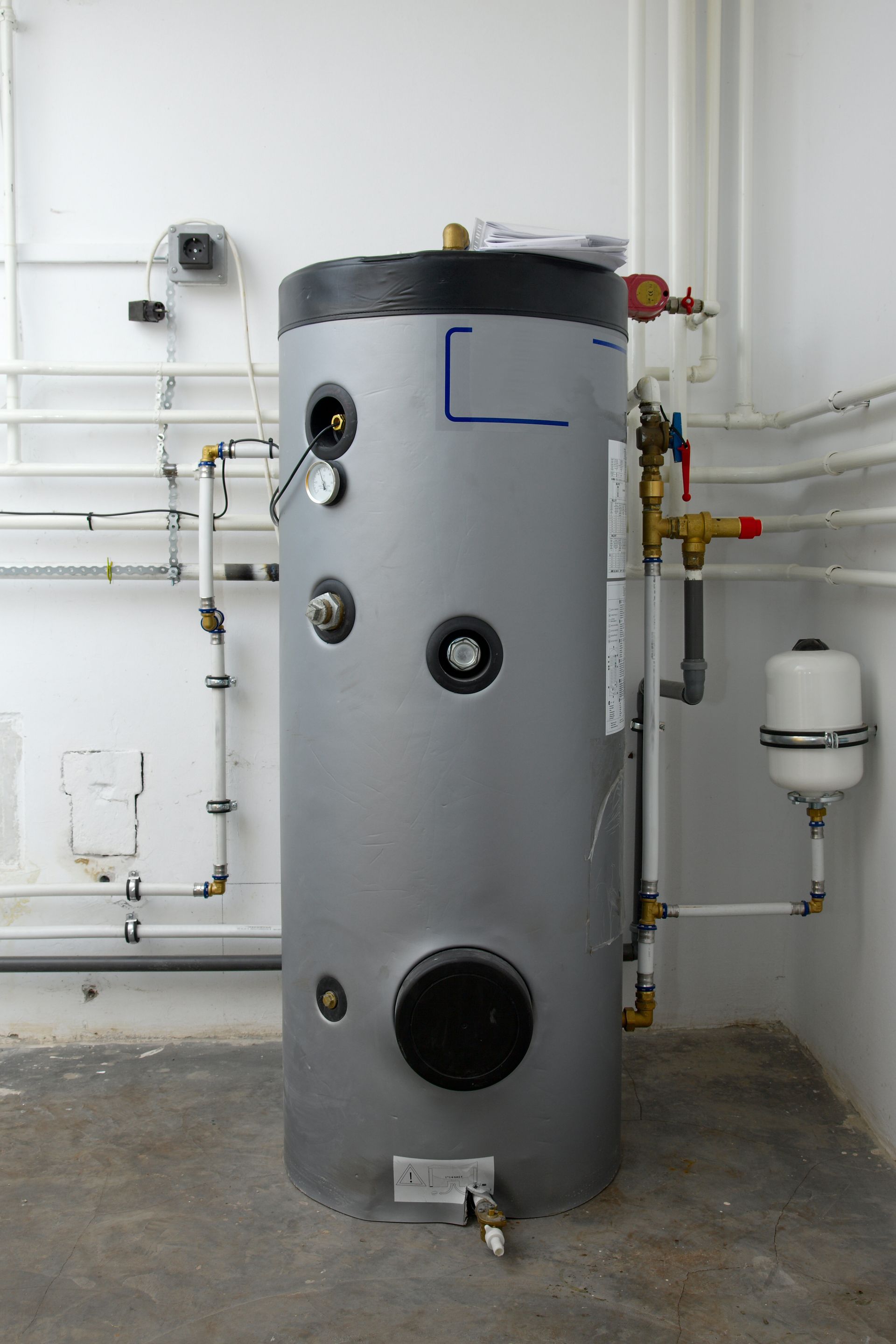 A grey hot water cylinder stands in a utility room, connected to white pipes and a small expansion tank on the wall.