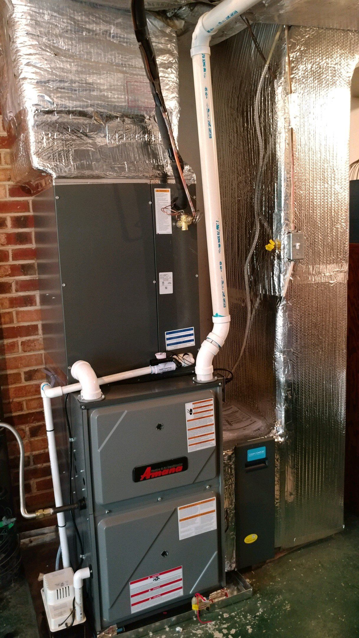 A furnace and HVAC system, with silver ductwork and white PVC pipes, is installed in a room with brick walls.
