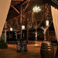 Dimly lit rustic hall with chandeliers, wooden beams, a chalkboard sign, and a barrel on the floor
