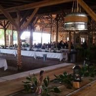 Rustic open-air dining hall with long tables, white tablecloths, and hanging lights beneath wooden beams.