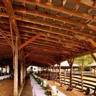 Rustic covered walkway with wooden beams, long white tables, and potted plants in a barn-like venue