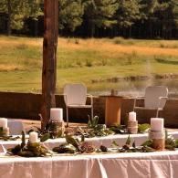 Outdoor dining table set with white chairs and candles beside a grassy field and trees