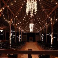 Indoor venue with warm string lights and chandeliers over a wooden aisle, decorated for an event.