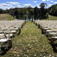 Outdoor wedding aisle lined with white chairs leading to a lakeside ceremony site under a sunny sky