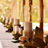 Row of white candles and greenery on a long outdoor table at a warm, sunlit event