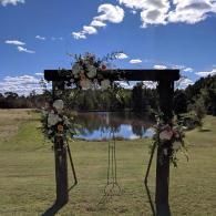 Flower-decorated wooden arch by a pond in a grassy field under a blue sky