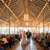 Wedding ceremony in a rustic barn, with bride and groom walking down the aisle between seated guests.