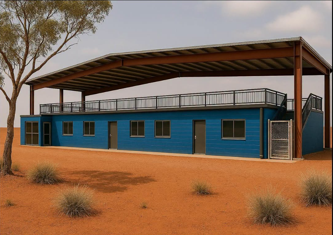 Blue desert building with a metal roof and tall posts, set in sandy terrain with sparse trees.