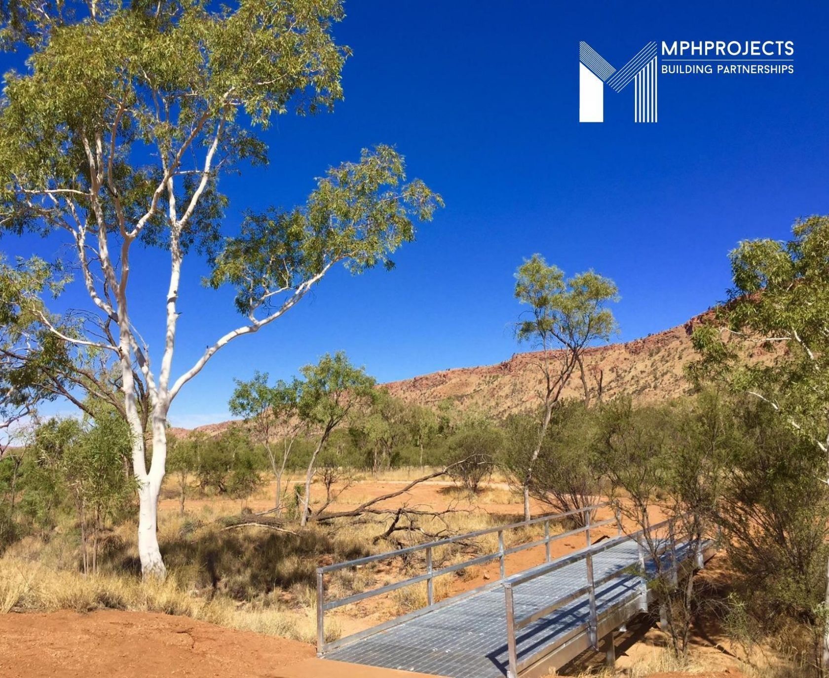 Metal bridge in a desert landscape with trees and mountains under a bright blue sky.