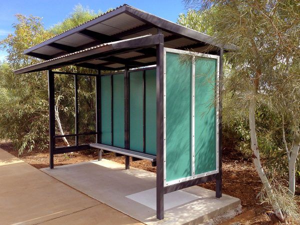 Bus shelter with black frame, green panels, and gray roof. Contains a bench, set on a concrete pad, and next to trees.
