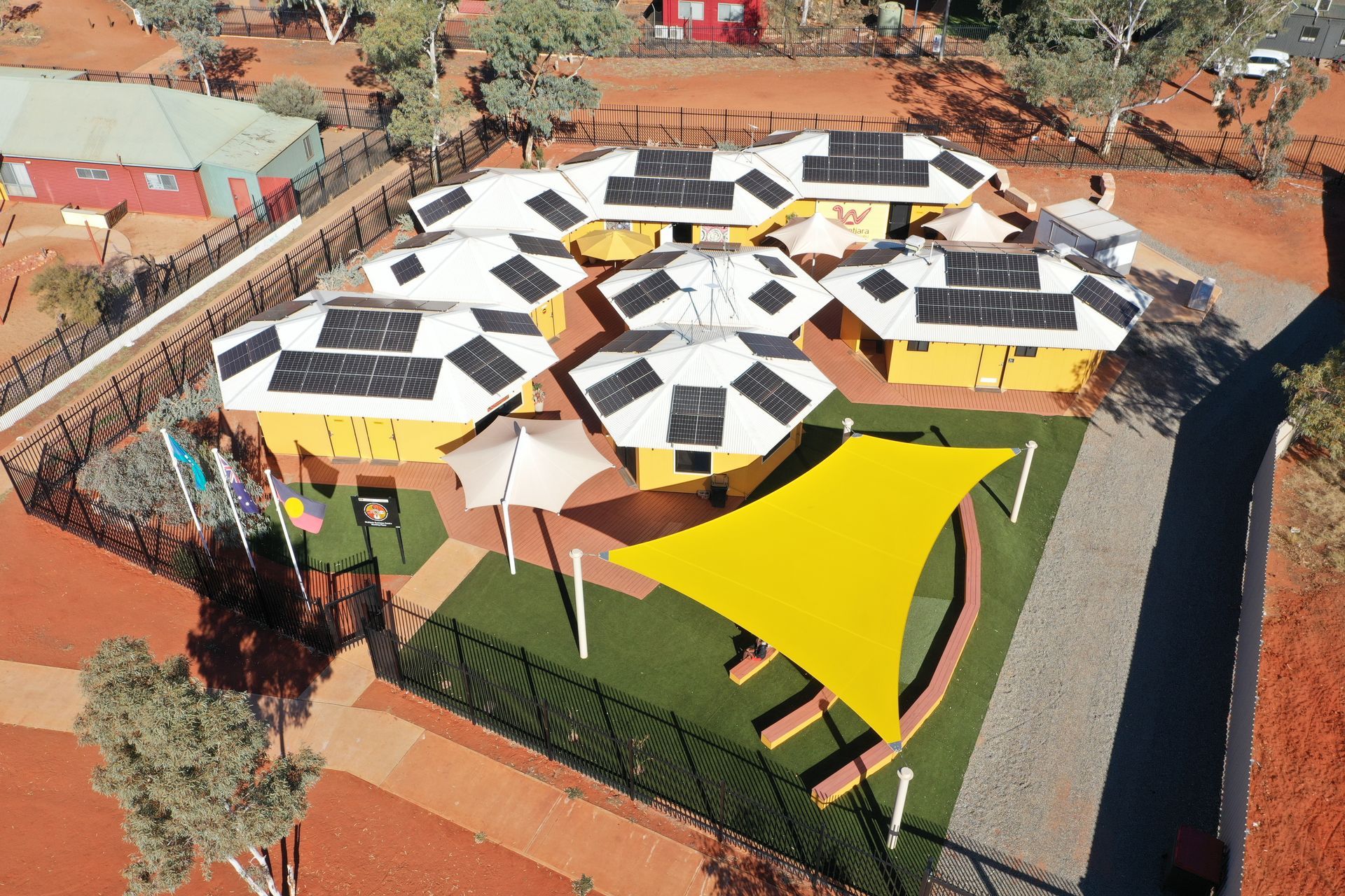 Aerial view of a yellow school building cluster with solar panels on multi-faceted roofs and a large yellow shade sail.