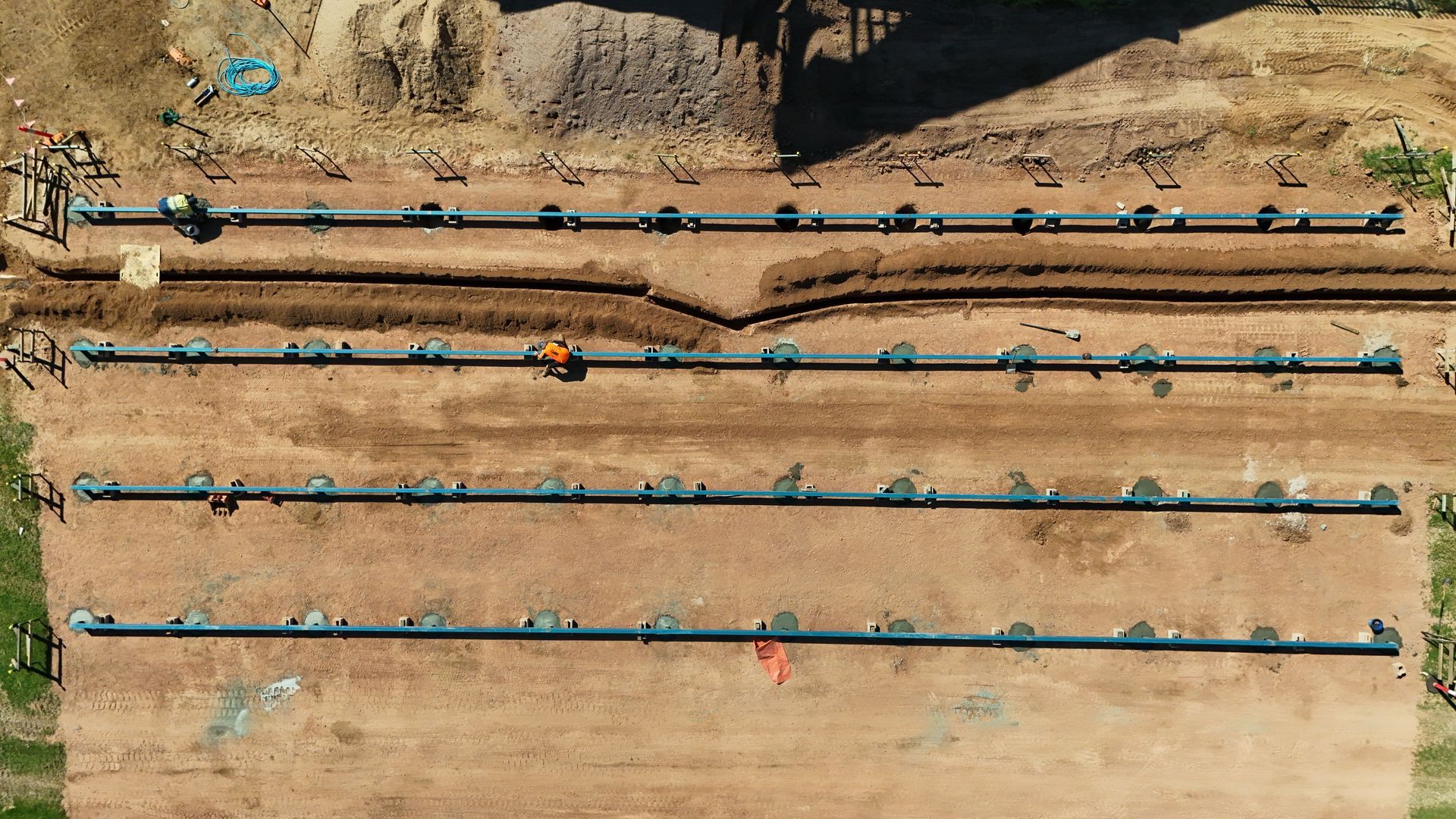 Aerial view of long irrigation pipes laid across a dry tan field, with small workers and equipment nearby