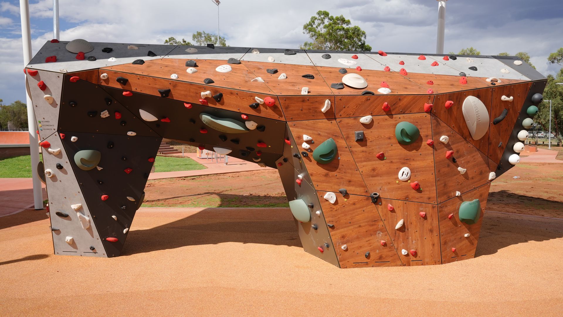 A wooden bouldering wall for children in a park, featuring a variety of colorful climbing grips and a gravel surface.