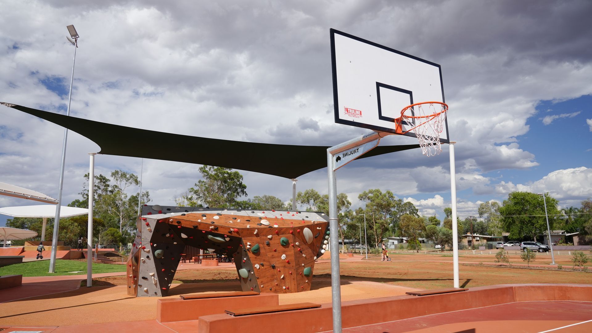 Basketball hoop and climbing wall in a park setting under a large shade structure; sunny day.