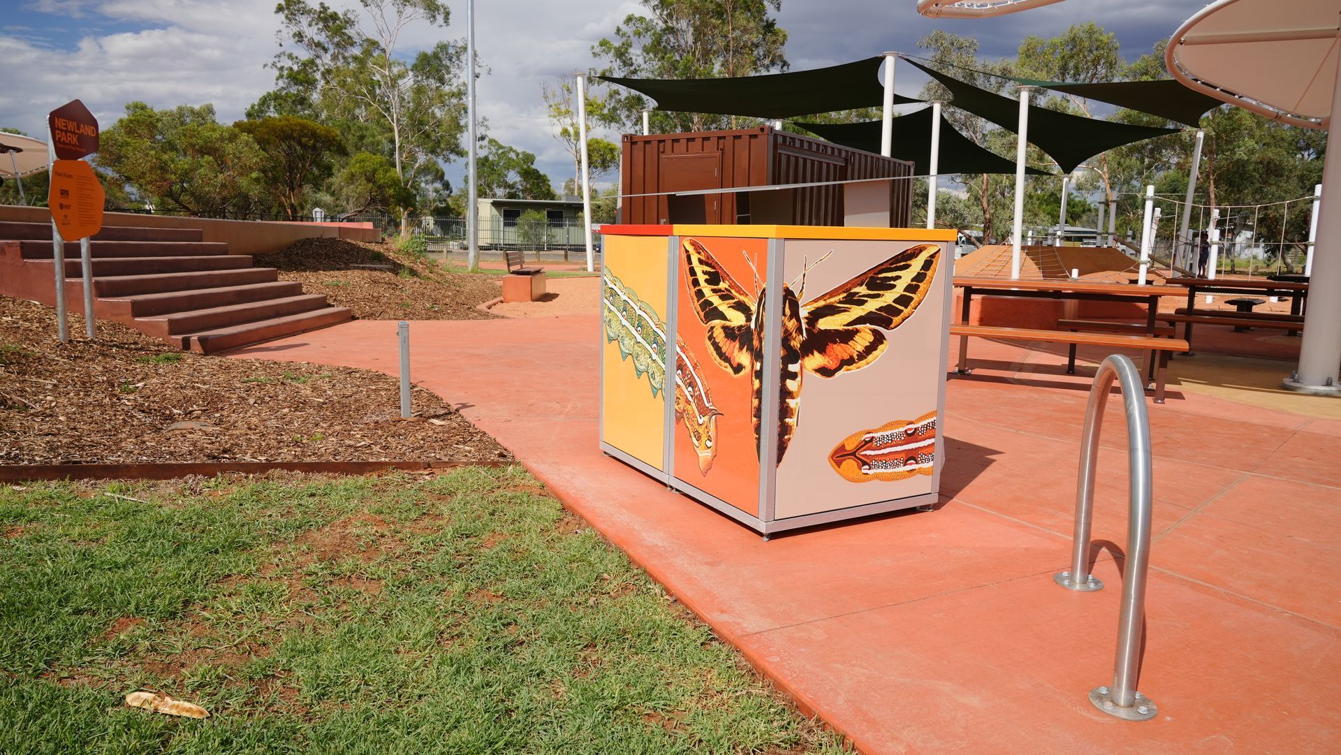 Colorful bin with butterfly art in a park setting with picnic tables and shade.
