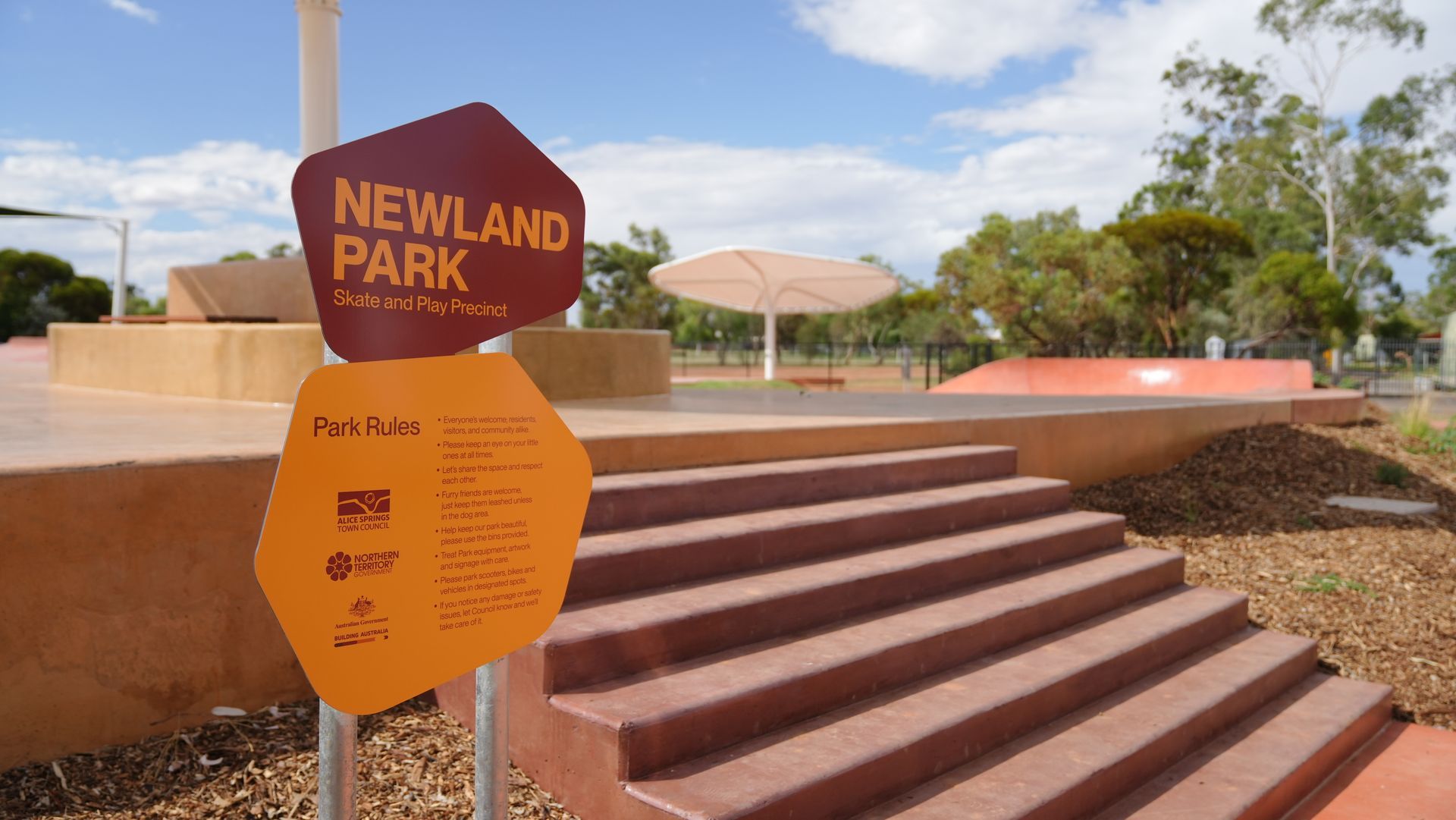 Sign for Newland Park, Australia. Orange sign with brown text, steps up to play area, blue sky.