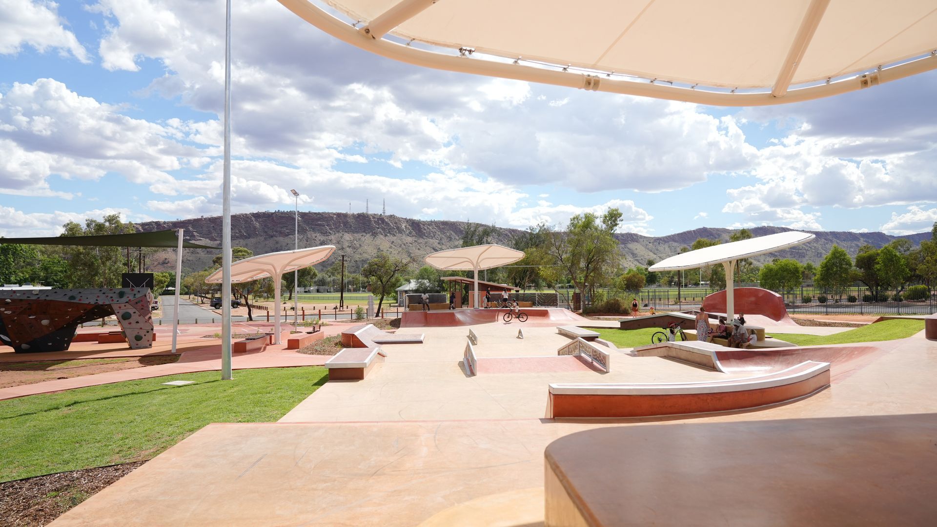 Skatepark with curved ramps, benches, and shade structures; mountains in the background, sunny day.