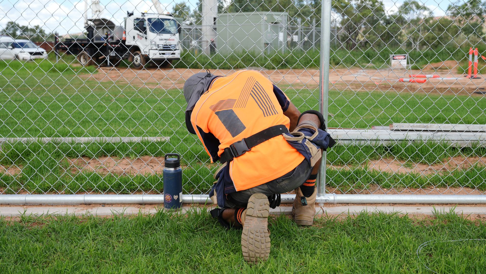Worker in orange safety vest kneels by a chain-link fence near a roadside ditch.