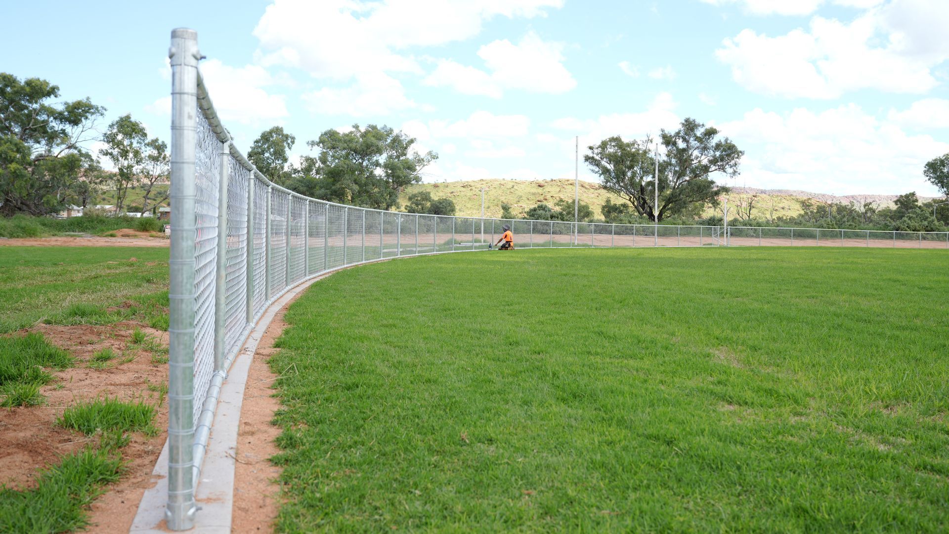 Curved chain-link fence bordering a green sports field with trees in the background