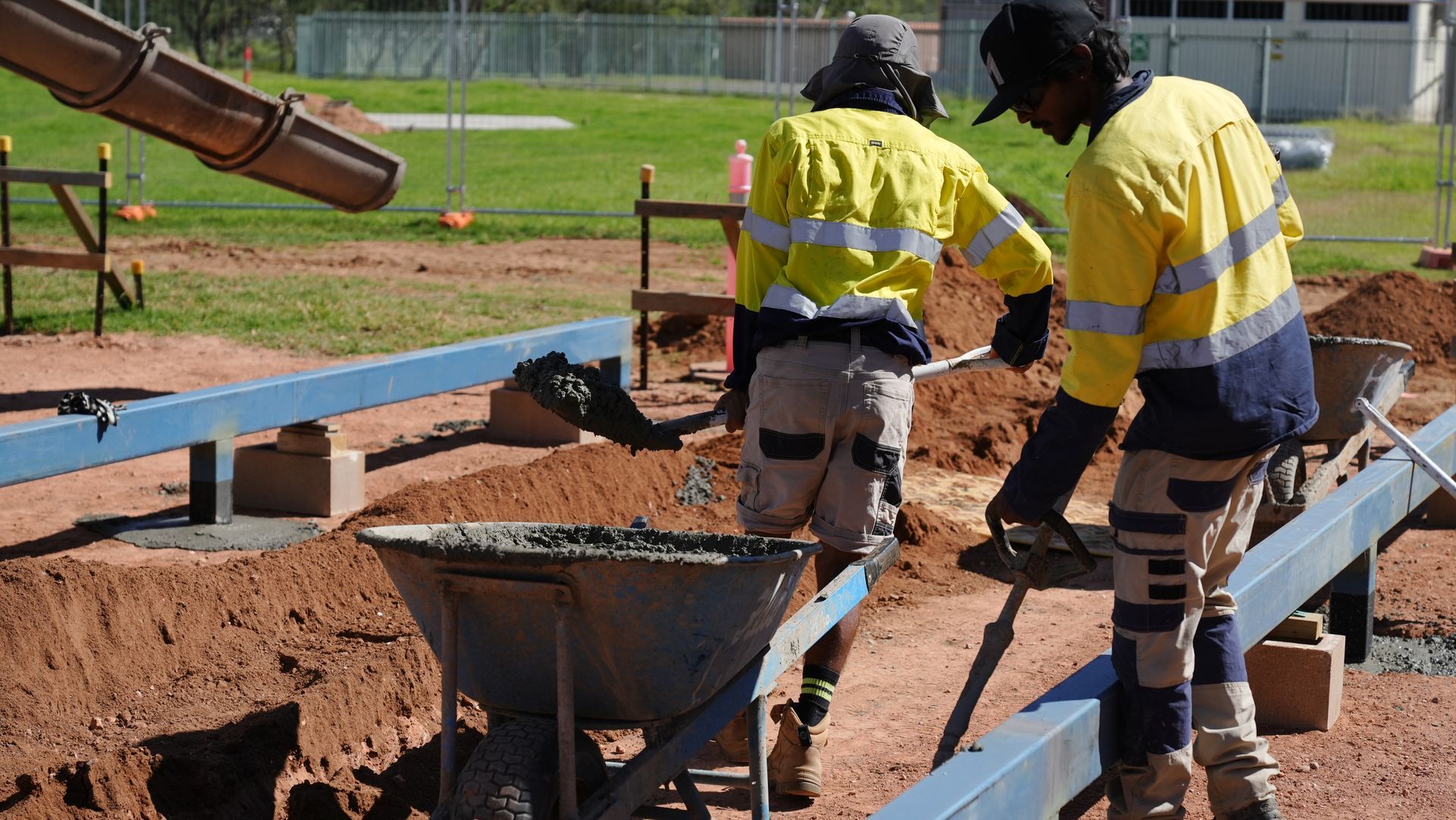 Workers in yellow safety vests pour concrete into a sidewalk mold at a construction site.