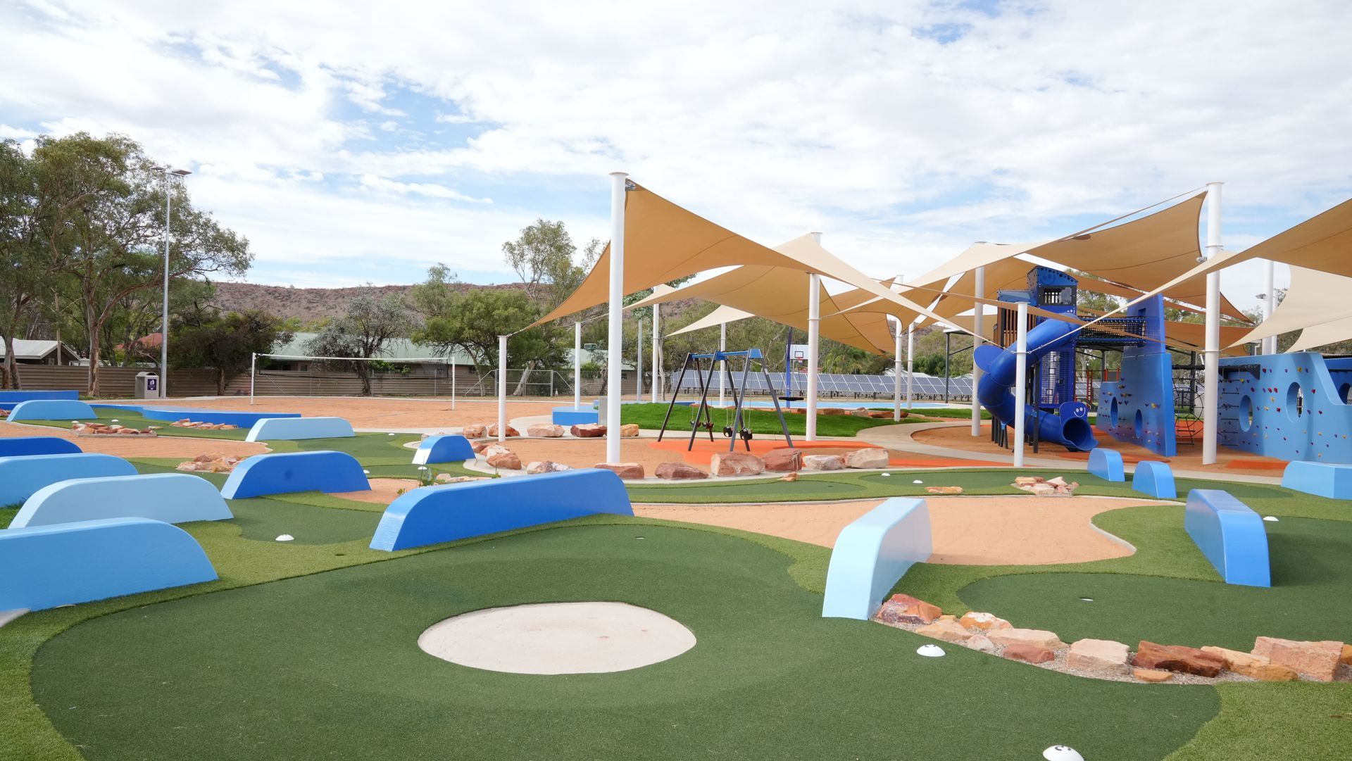 Playground with blue and tan structures under shade sails, green turf, and tan ground.