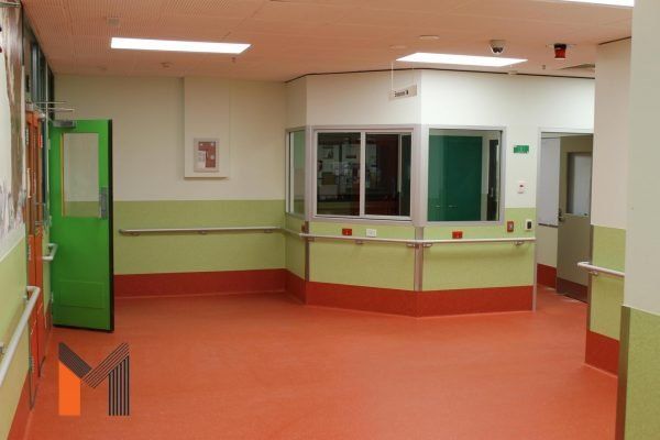 Hospital hallway with orange floor, green and white walls, and a glass-walled nurses' station.