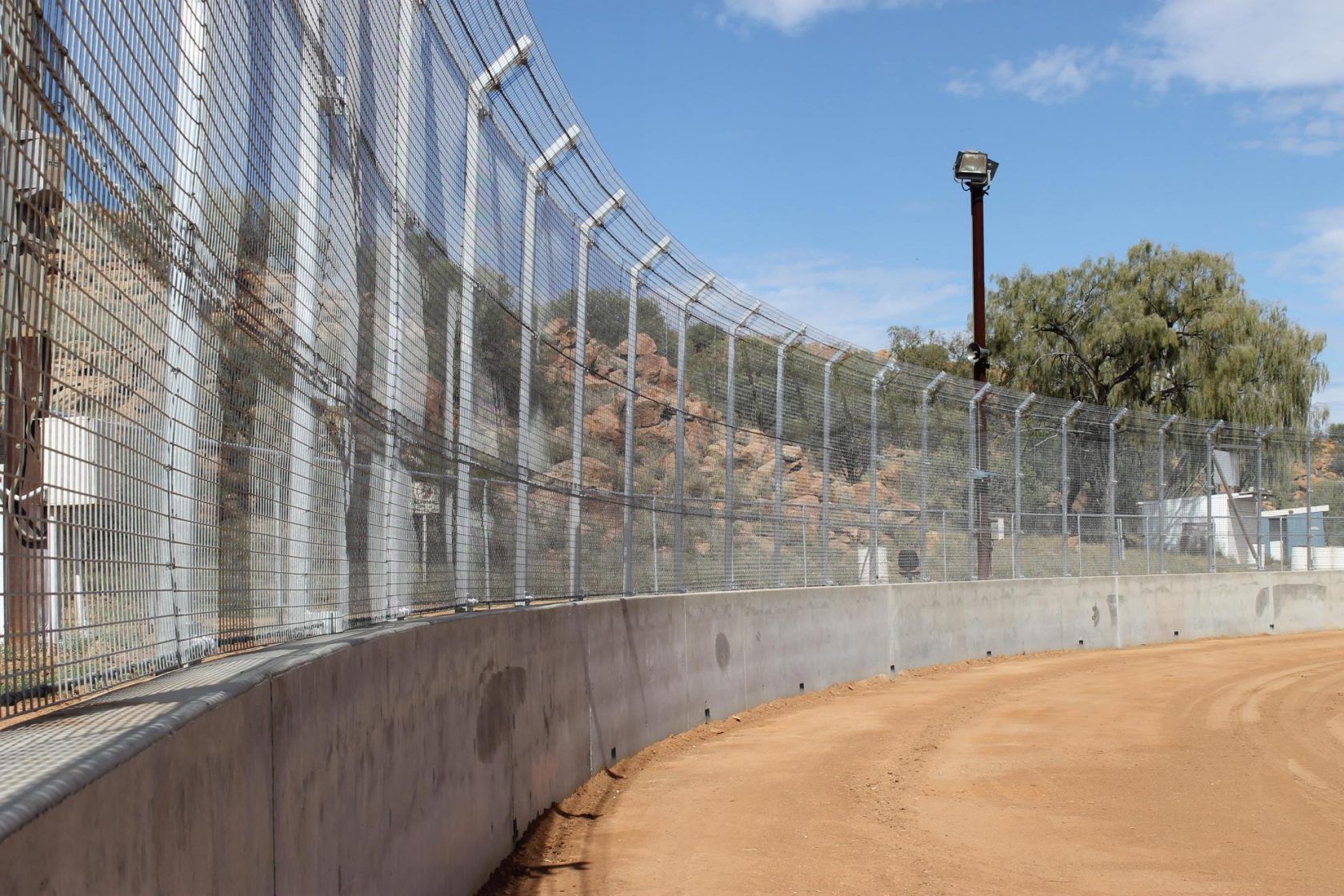 A curved dirt race track with a concrete barrier and chain-link fence, under a partly cloudy sky.