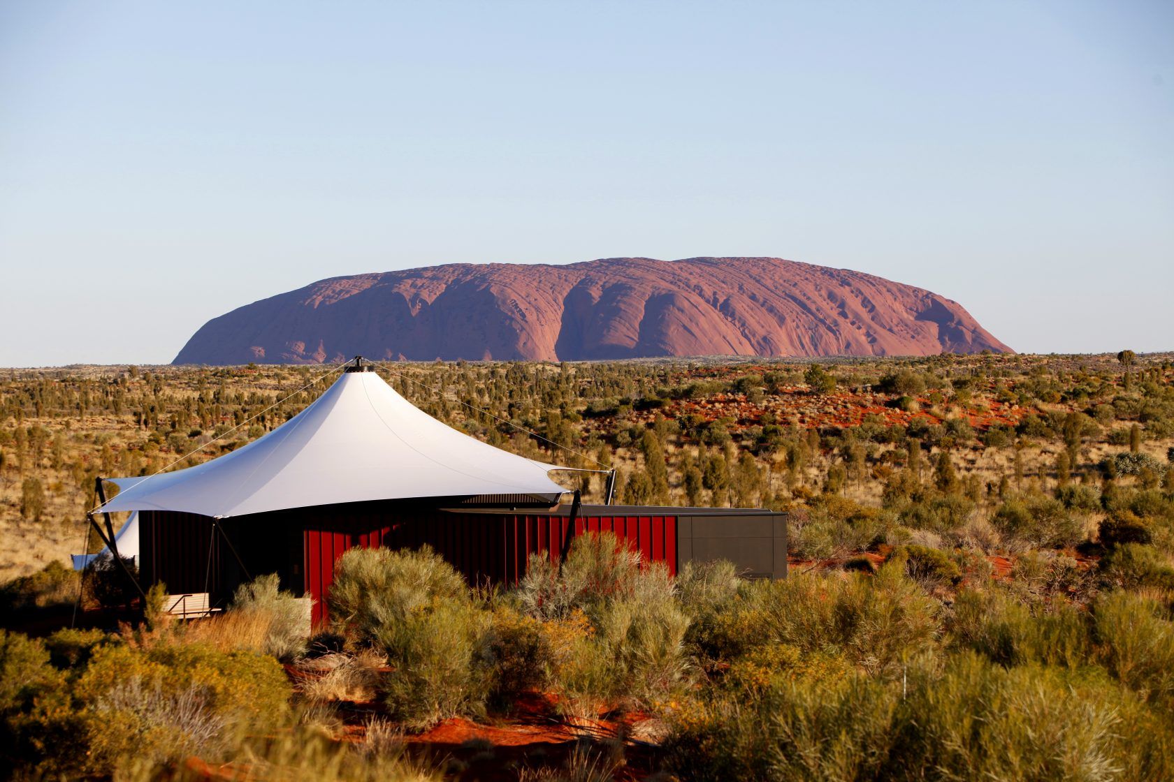 A tent with Uluru in the background, set in a desert landscape with red earth and green bushes.