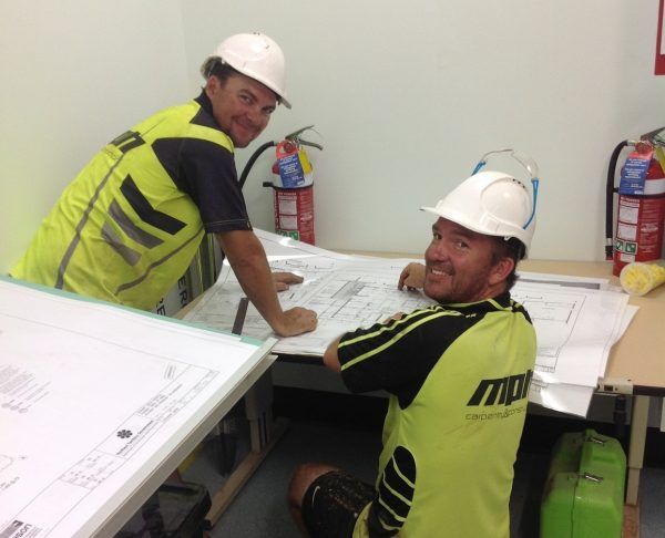 Two construction workers in white helmets, examining blueprints in an office setting. They are smiling.