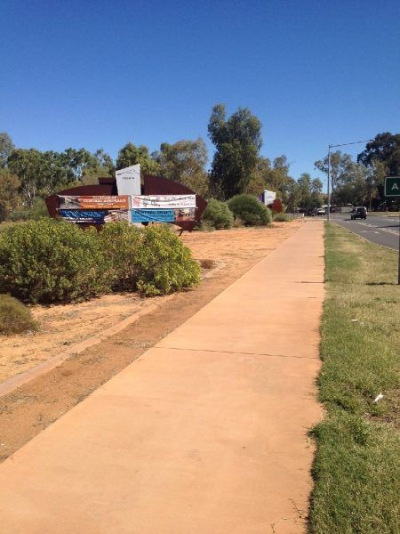 Sidewalk alongside a road and low bushes. Building with signs and trees in background on sunny day.