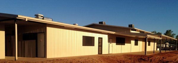 Low-angle view of a beige building with a long porch, brown dirt, and a clear blue sky.