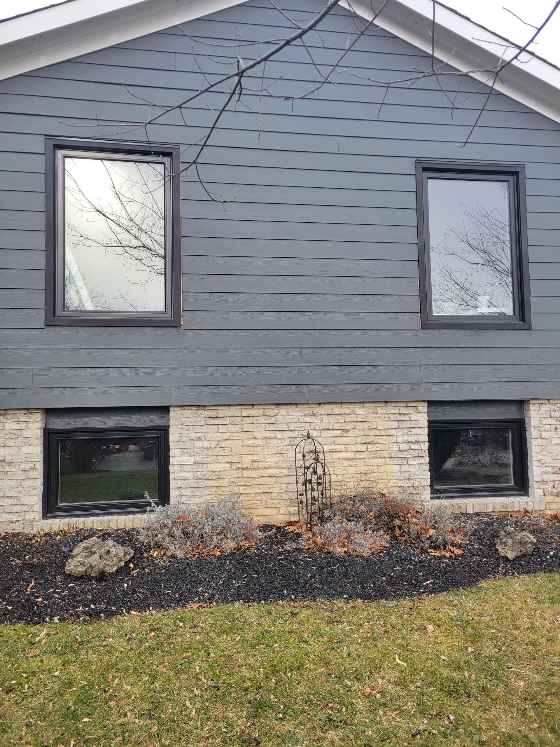 Gray house with four dark-framed windows; two above, two below. Tan brick base. Brown and black mulch, dead plants.