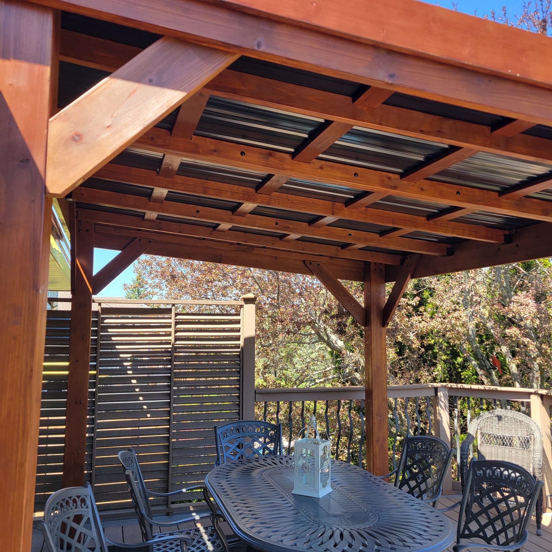 Wooden pergola over patio with a metal roof. A table and chairs are beneath.