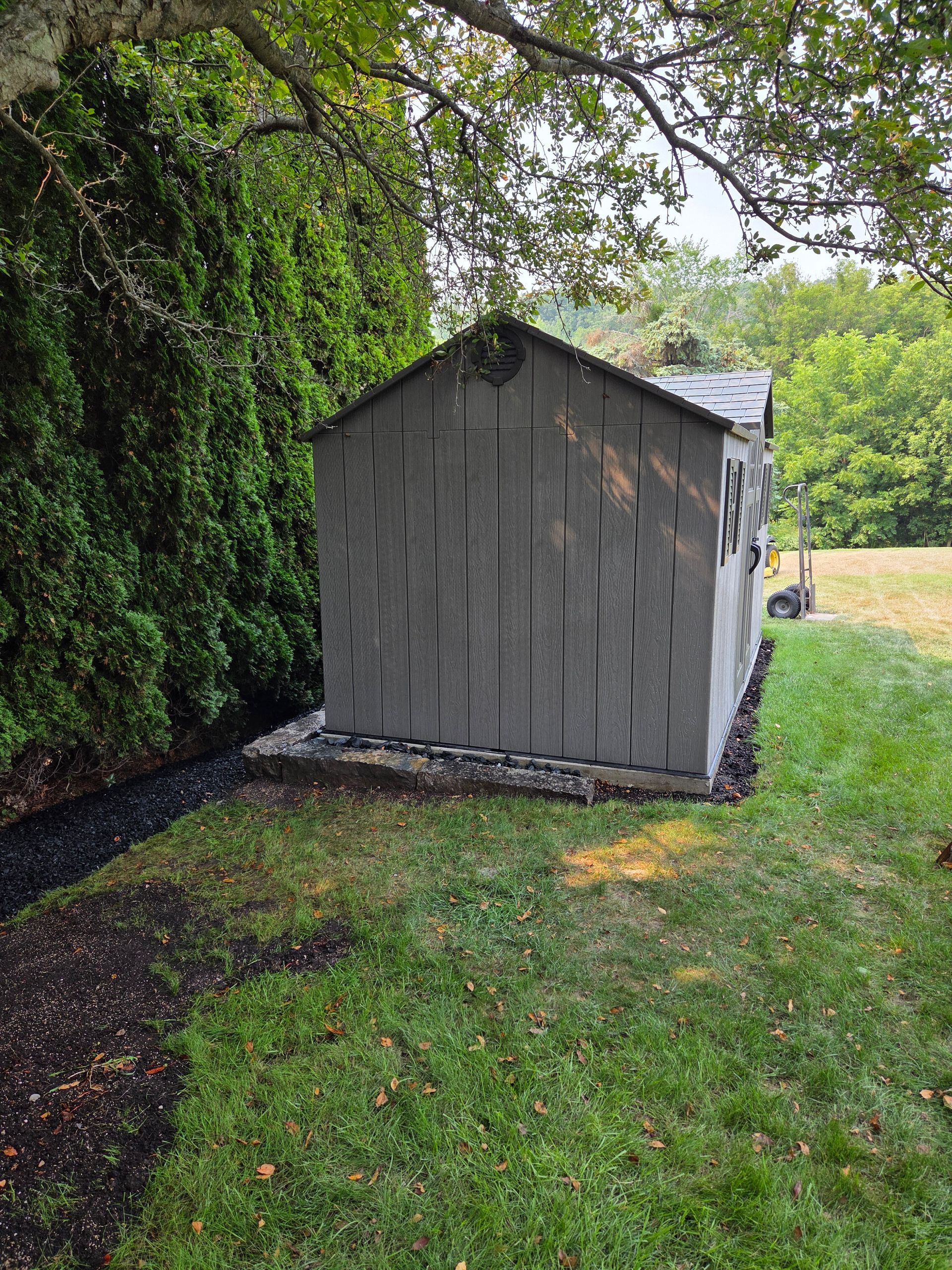 A gray shed with a dark roof stands on a concrete foundation in a grassy yard, beside a hedge.