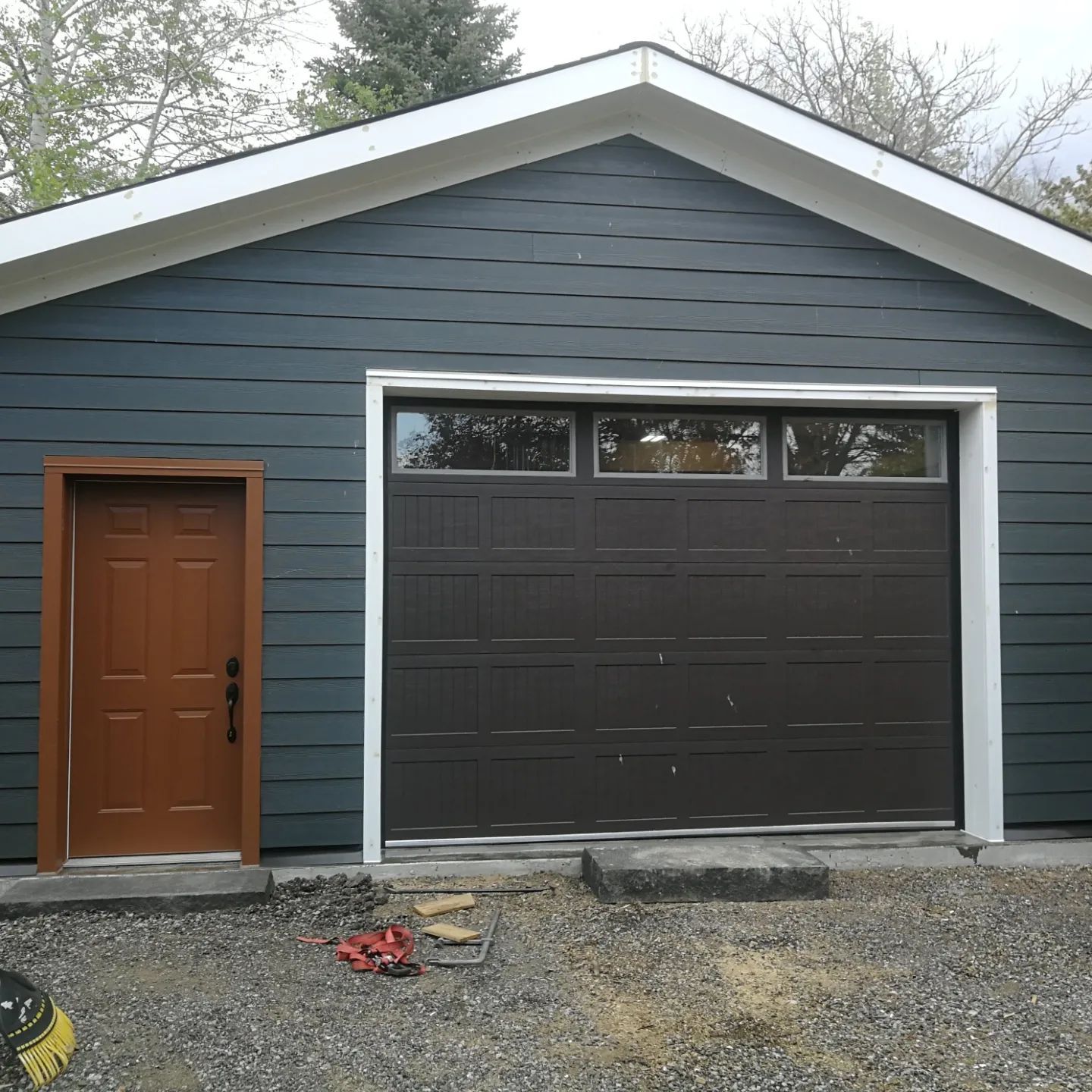 Garage exterior with blue-gray siding, brown door and garage door, white trim, and gravel driveway.