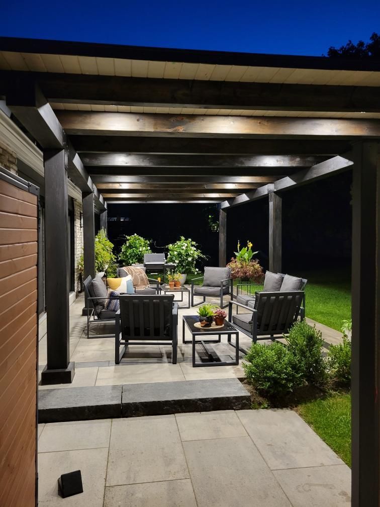 Patio with dark furniture under a wooden pergola, illuminated at night. Lush plants and stone flooring.