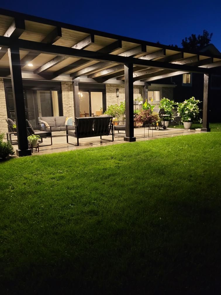 Night view of a patio with pergola, seating, and potted plants, illuminated by overhead lights and the glow of the house.