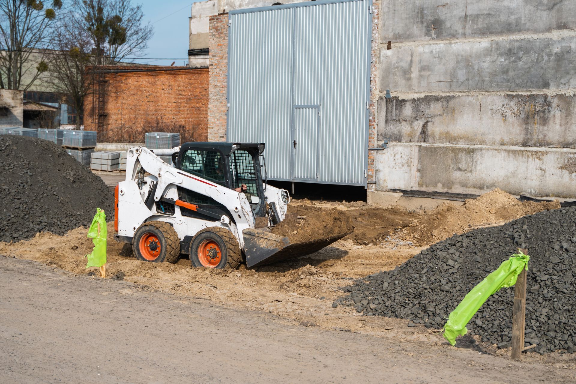 Bobcat loader moving dirt at a construction site beside a metal shed and concrete wall