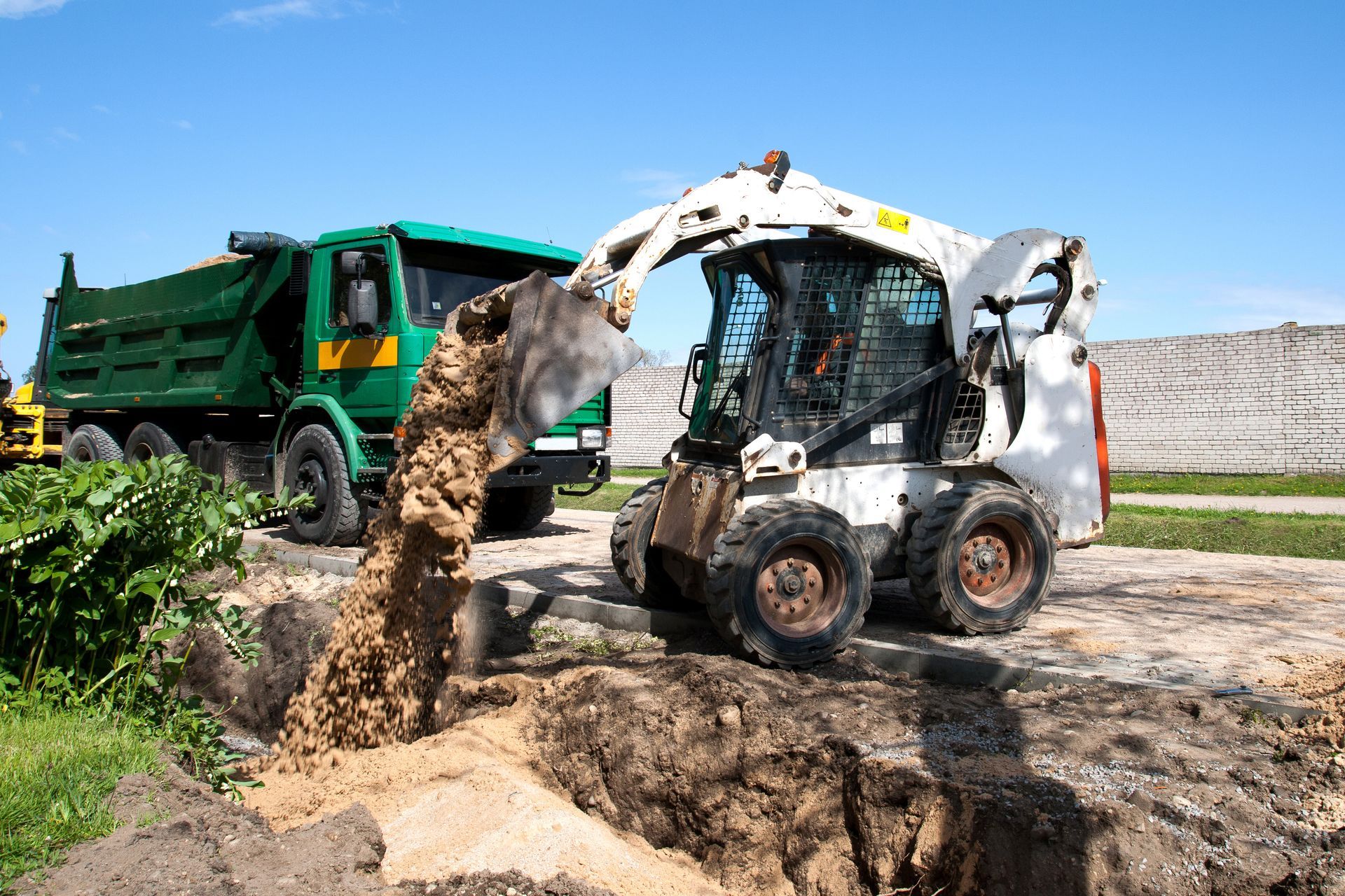 Bobcat dumping dirt into a trench beside a green dump truck on a construction site