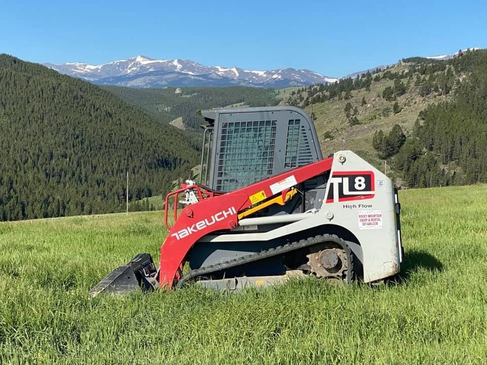 White and red Kubota skid steer in a grassy mountain field with snowcapped peaks in the background