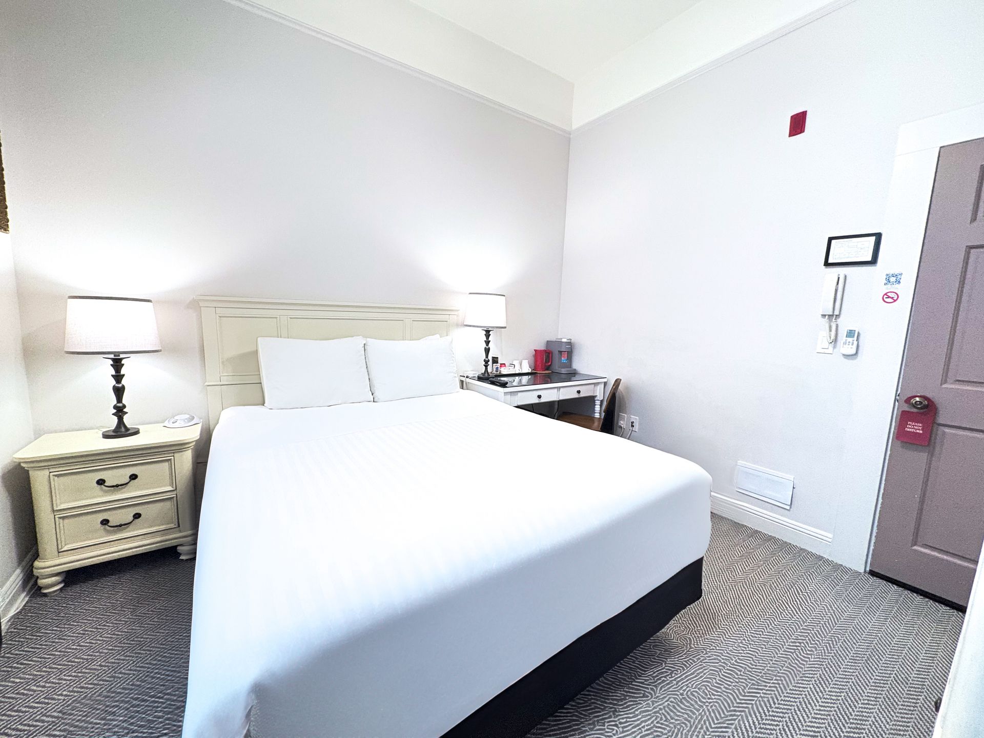 A hotel room with a bed , nightstand , lamp and door.