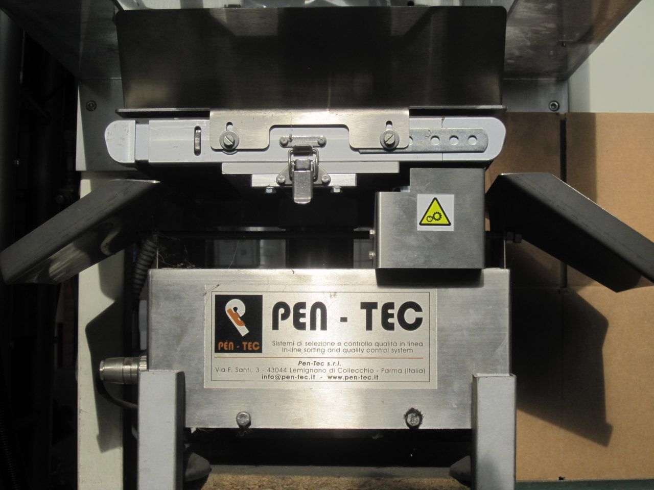 Primo piano di una macchina industriale PEN-TEC con componenti metallici, etichetta di avvertenza e meccanismo centrale.