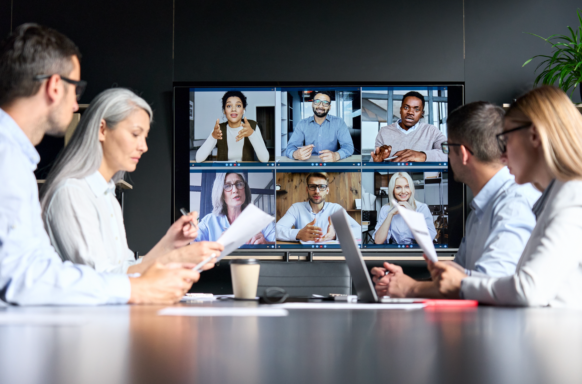 People in a conference room participate in a video call on a large screen, collaborating with other individuals remotely.