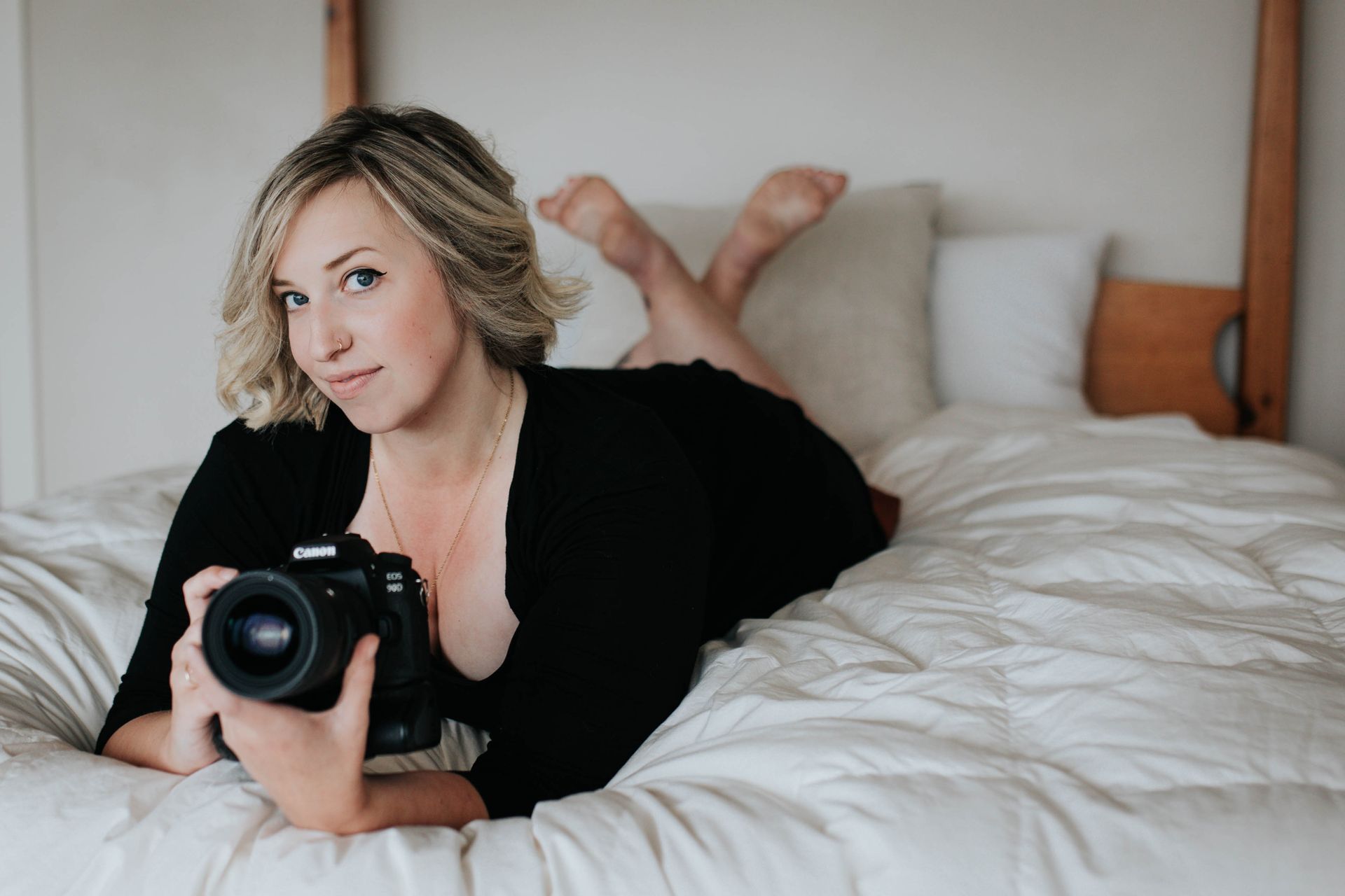 Blonde female holding DSLR camera while posing on bed