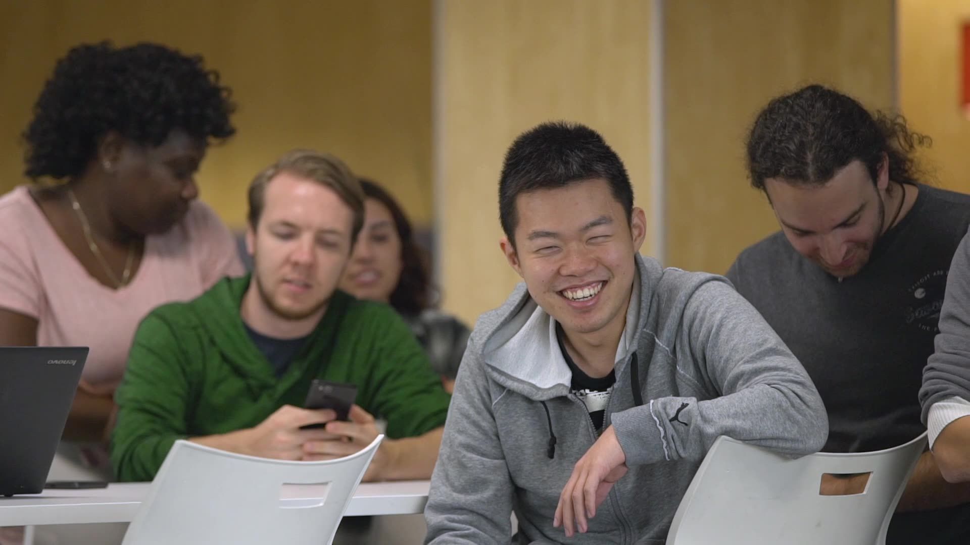 People seated at laptops in a meeting, smiling and talking in a classroom or office setting.