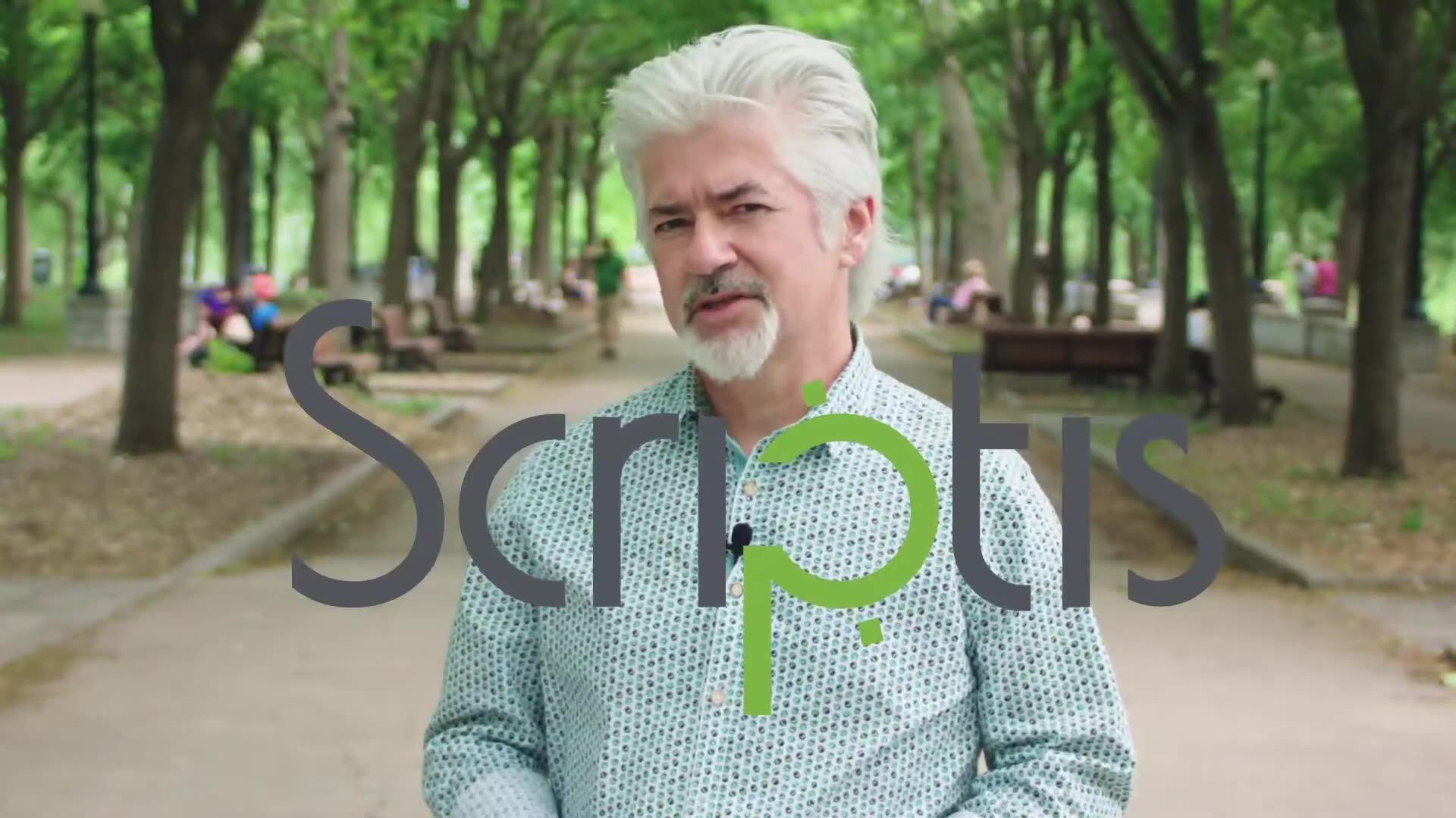 Man with white hair and beard sitting in a park with trees and a path behind him