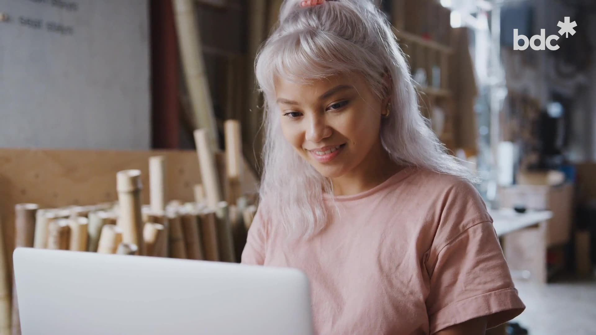 A person with light hair in a pink shirt works at a laptop in a workspace filled with stacks of wooden poles.