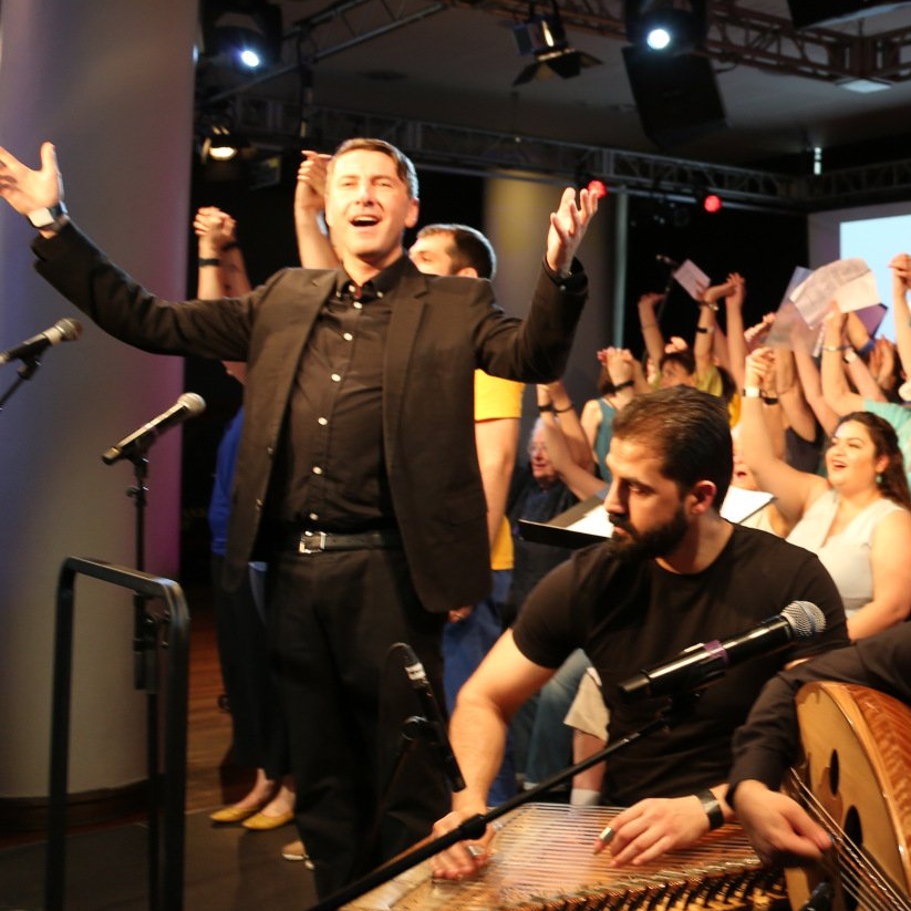 A man in a black suit is standing in front of a crowd with his arms in the air